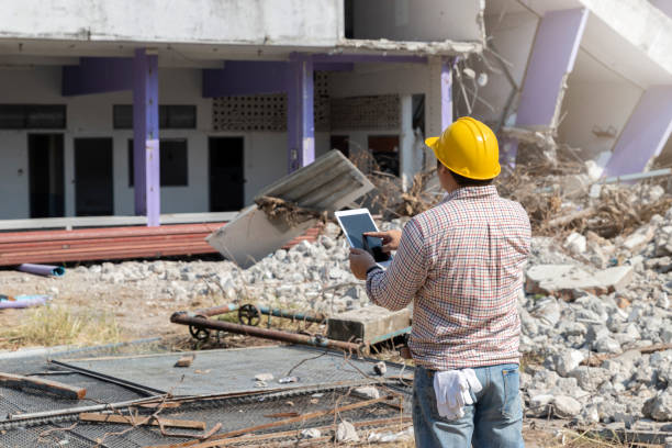 Construction worker in a yellow hard hat inspecting a damaged building site with rubble and broken flooring.