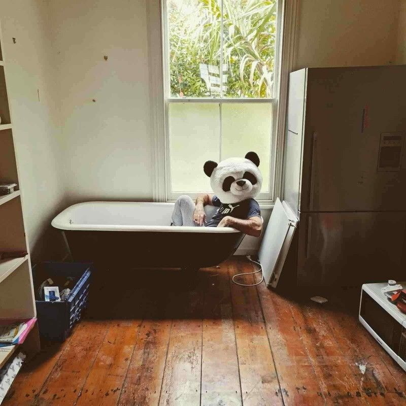 Person wearing a panda head costume sitting in a bathtub placed in the middle of a room with Engineered wooden flooring, refrigerator, and shelves.