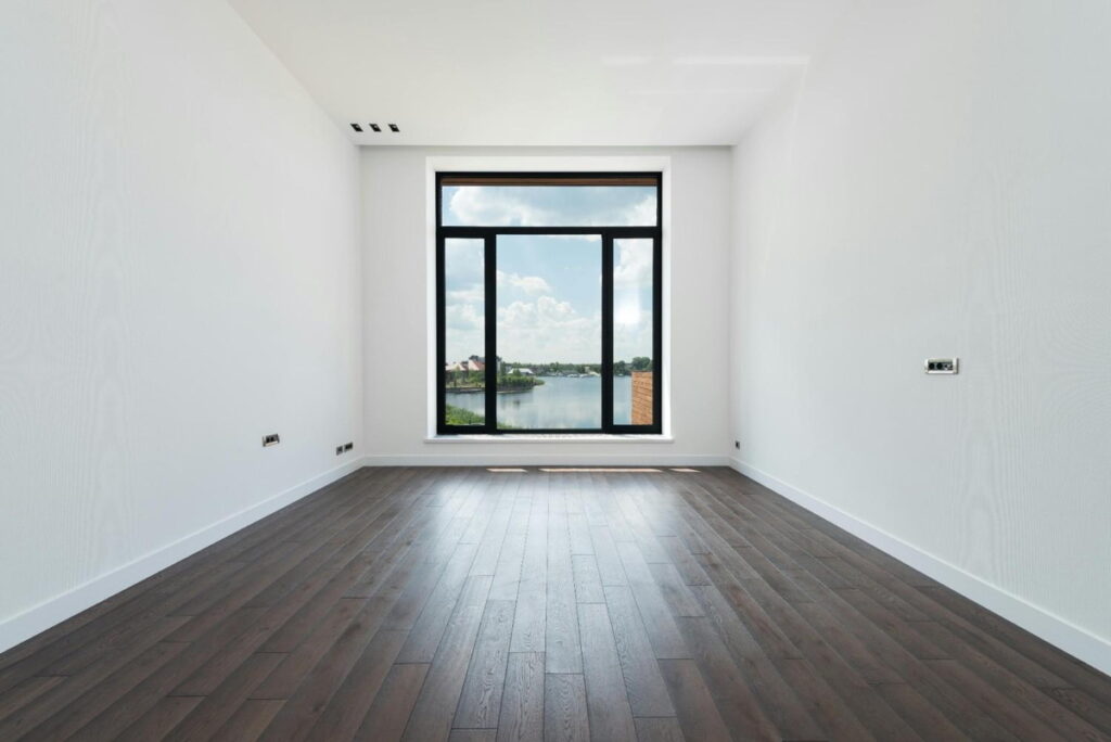 Empty room with dark brown laminate wood flooring, white walls, and a large window showing a lake and sky view.