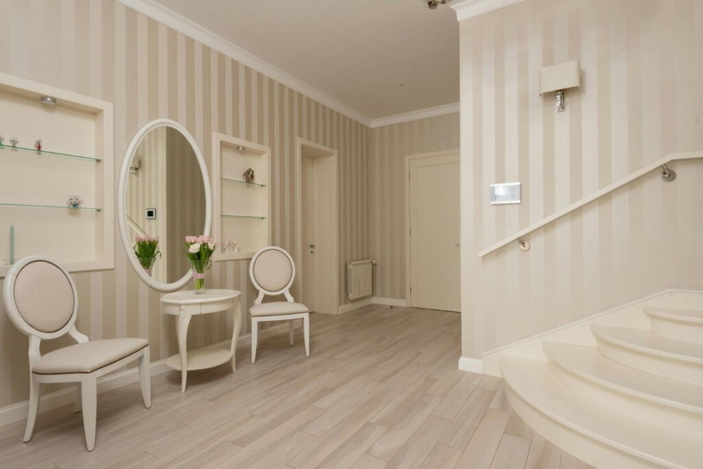 allway interior with light oak laminate wood flooring, striped beige wallpaper, two white chairs, round table with flowers, and oval mirror.