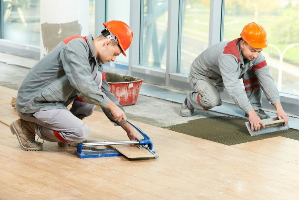 wo workers in helmets and uniforms repairing flooring; one cutting a wooden plank with a manual cutter, the other applying adhesive and fitting planks on the floor.