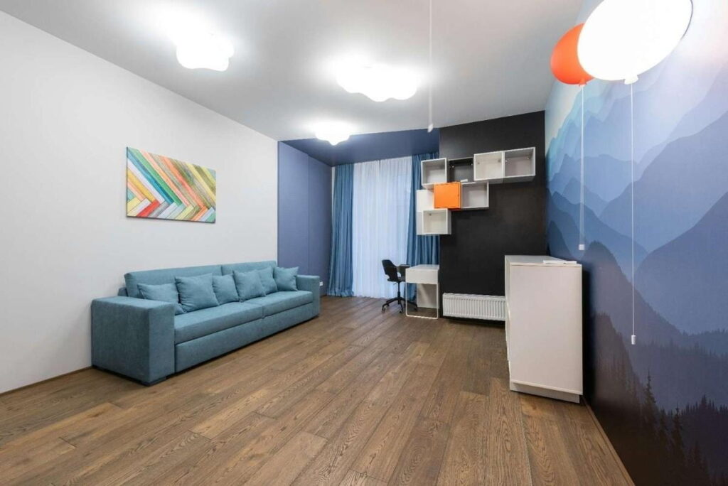 Living room interior with dark oak laminate wood flooring, teal sofa, wall shelves, study desk, colorful wall art, and mountain mural accent wall.