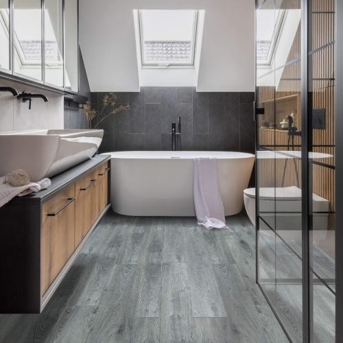 Bathroom interior with grey laminate wood flooring, freestanding bathtub, dual vessel sinks on wooden vanity, glass shower enclosure, skylights, and black fixtures.