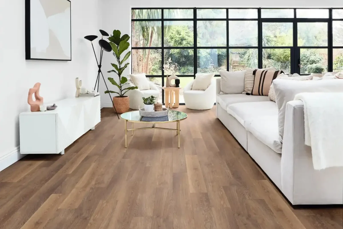 Living room with warm oak luxury vinyl plank flooring, white sectional sofa, glass coffee table, indoor plants, and large black-framed windows letting in natural light.