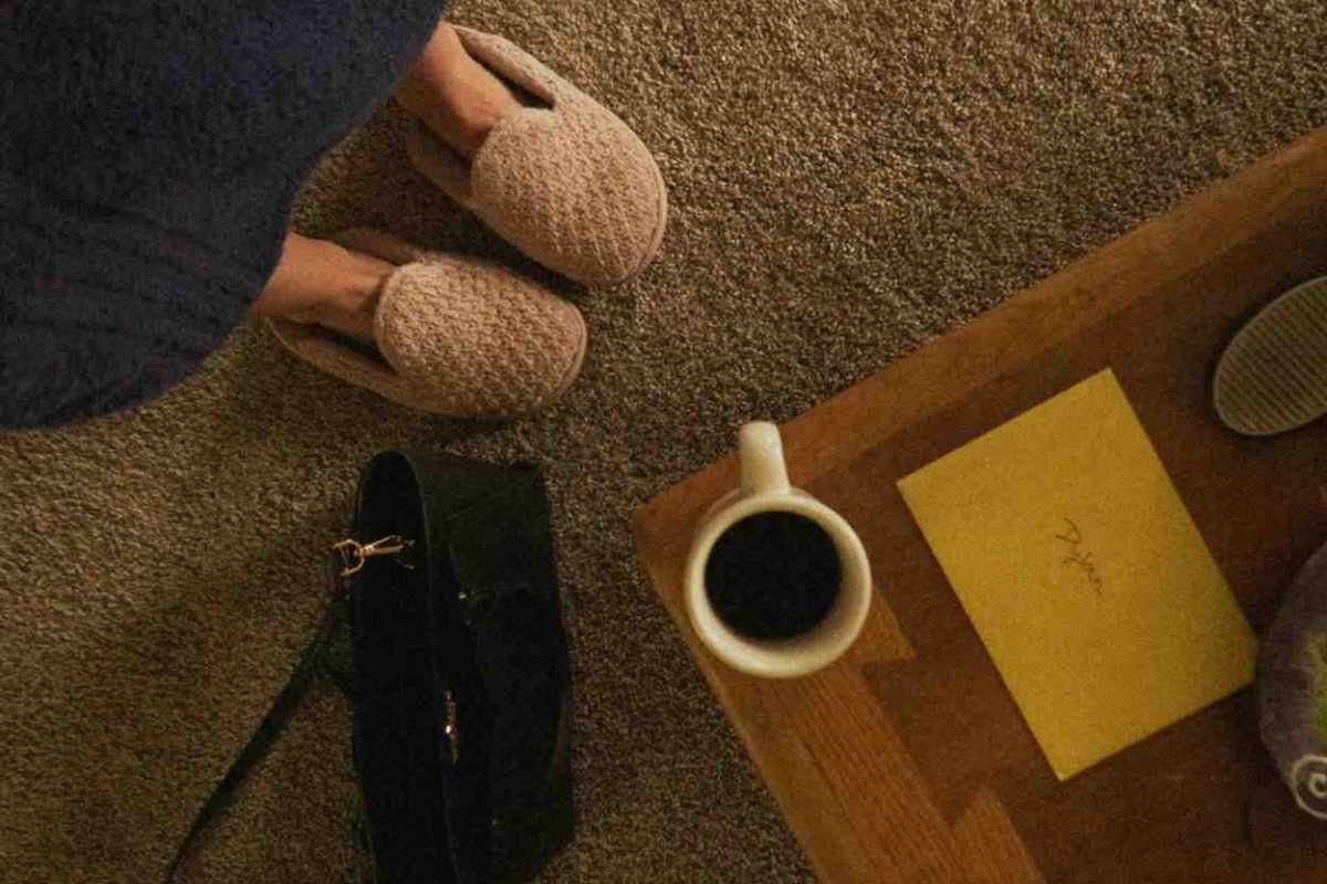 Beige carpeted floor with a wooden coffee table holding a coffee mug and yellow envelope, black handbag on the floor, and a person wearing slippers and a robe standing nearby.